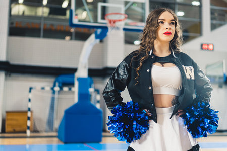 Horizontal shot of cheerleader wearing red lipstick in a jacket posing with blue shiny pom-poms....