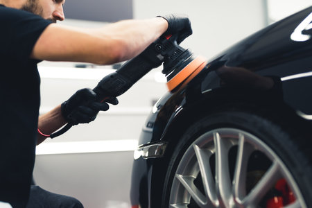 Low angle view of professional car detailing male worker using orbital polisher to polish black car...