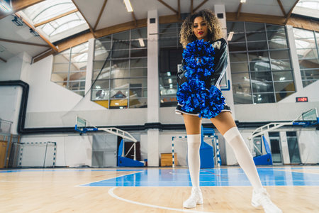 Full shot of cheerleader with curly hair posing with blue shiny pom-poms. Basketball court blurred in the background. High quality photoの写真素材