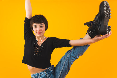 Flexible female dancer showing her skills by stretching one leg up and looking at camera. Casually dressed female yoga instructor stretching one leg in the air. Yellow background. High quality photoの写真素材