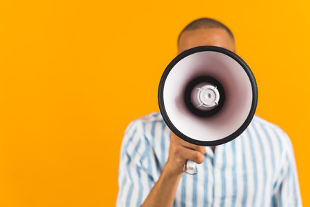 Young Black male announcer covering his face with bullhorn over orange background. Medium studio shot of man holding megaphone. High quality photoの写真素材