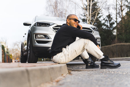 Stylish young adult man in sunglasses sitting on pavement in front of his expensive car and having a phone call. Outdoor full-length shot. High quality photoの写真素材