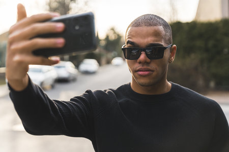 Confident casually-dressed short-haired man in sunglasses taking selfie with modern smartphone outdoors. Blurred background. High quality photoの写真素材