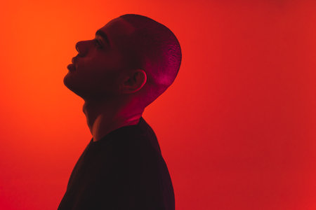 Thinking about problems. Side portrait of Black young adult man looking up and bending his neck. Studio portrait over neon read background. High quality photoの写真素材