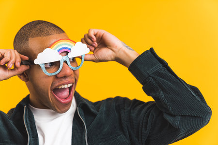 Excited African American man wearing paper party glasses with rainbow and clouds on yellow...
