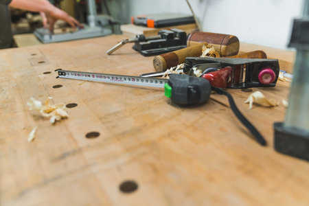 Measure tape, mallet, wood filings, and other necessary tools used carpentry lying on wooden table. Carpenter hands in the background. High quality photoの写真素材