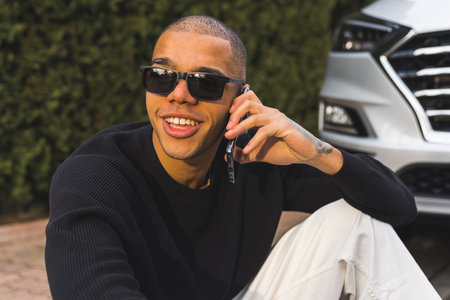 Young smiling man in sunglasses sitting on a curb near the car and speaking on the phone. High quality photoの写真素材
