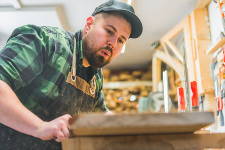 young male carpenter cleaning the wood by blowing sawdust on a piece of wood, wood products. High quality photoの写真素材