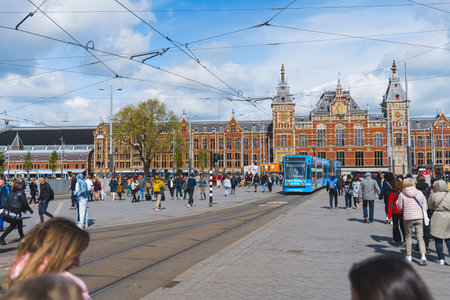 Amsterdam Centraal Station photographed during sunny day. Blue tram going in the middle. Groups of tourists in light jackets walking by. High quality photoの写真素材
