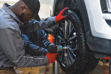 Two men auto mechanics changing car tire on a vehicle on a hoist using an electric drill to loosen...