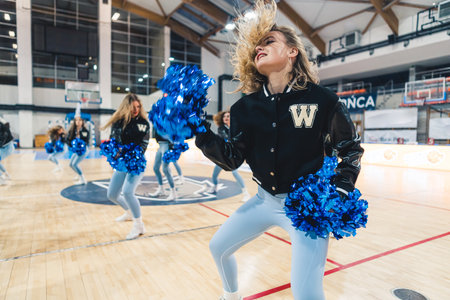 Dynamic shot of college girls cheerleaders in uniforms dancing with pompons on a basketball court. High quality photoの写真素材