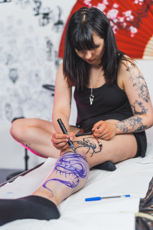 tattooed girl sitting on the table and drawing with a marker on her leg, Freehand technique. High quality photoの写真素材