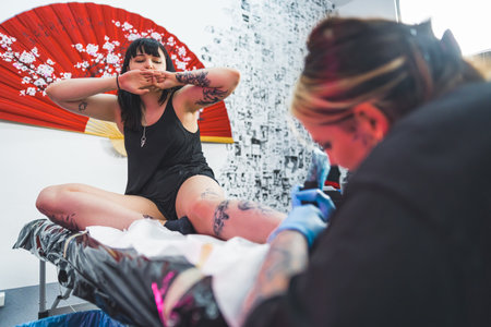 tattooist girl making a new tattoo on a customers leg sitting on the table, full shot. High quality photoの写真素材