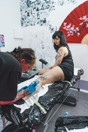 vertical shot of a girl tattooist drawing a tattoo on a girls leg, working in the tattoo salon. High quality photoの写真素材
