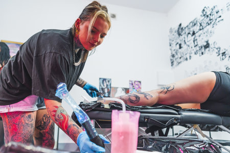 Caucasian young tattooist girl drawing a tattoo on a customers leg, medium full shot tattoo salon. High quality photoの写真素材