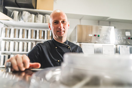 Closeup indoors shot of professional kitchen with stainless steel equipment and chef in black uniform. Bald caucasian man at work looking straight into camera. High quality photoの写真素材
