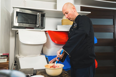 Medium indoors shot of bald caucasian man wearing black professional chef attire mixing ruby chocolate batter with whisk in kitchen. Professional chef at work in industrial kitchen next to microwave and kitchen cabinets. High quality photoの写真素材