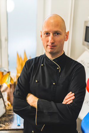Vertical portrait shot of profesional chef in black uniform. Caucasian bald male kitchen worker with confectionery appliances in background. Job description concept. High quality photoの写真素材