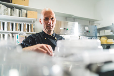 Cook in black apron smiling over detail shot of professional cooking machine in foreground and. Bald caucasian male chef enjoying work standing in kitchen. High quality photoの写真素材