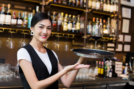 Young Asian Woman Waiter Restaurant Catering Service Happy Emotion. Woman Waiter Present Red-Wine for Customer at Bar. Woman with Wine in Bar Concept.の写真素材