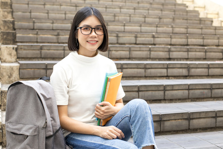 Portrait of Attractive Asian Student Woman sitting at outdoor place. Woman holding Book with...