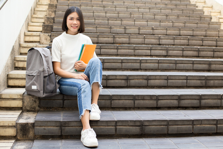 Portrait of Attractive Asian Student Woman sitting at outdoor place. Woman holding Book with Attractive Smiling to Camera. People with Education Concept.の写真素材