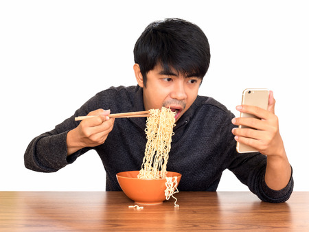 Man eating chinese noodle monstrously whilst looking and using smartphone isolate on white...