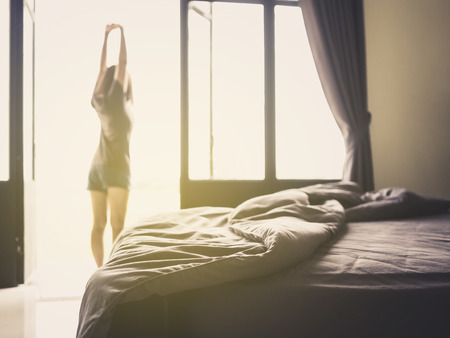 Confusion duvet and bed in luxury bedroom and healthy young woman stretching and relaxing by the window after waking up in the morning of holiday. Selective focus.の写真素材