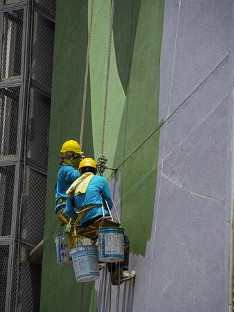 Chiang Rai, Thailand: September 18, 2019 - Two painters are painting the exterior of the building on a dangerous looking scaffolding hanging from a tall building.のeditorial素材