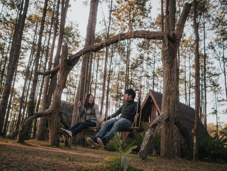 Young Asian man and woman lover playing swing in front of European-style resort wooden house or wooden hut with fun and happiness in a pine forest. The environment is similar to Europe or Canada.の写真素材