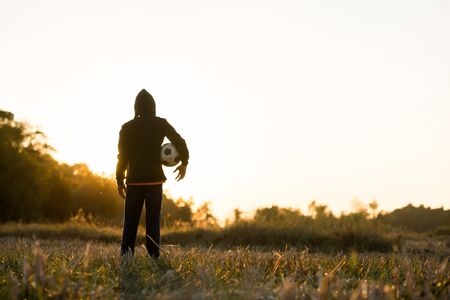 Asian boy playing football soccer in the meadow with sunset background