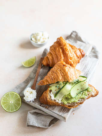Croissant sandwich with ricotta or cream cheese, mashed avocado and cucumber on wooden cutting board. Delicious brunch or breakfast. Selective focus.の写真素材