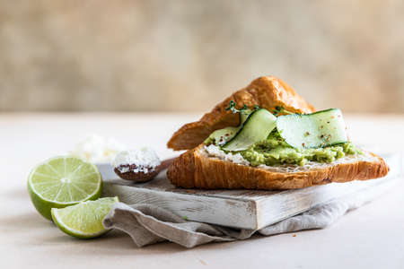 Croissant sandwich with ricotta or cream cheese, mashed avocado and cucumber on wooden cutting...