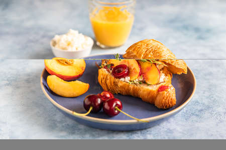 Fresh croissant with cream cheese, nectarine and cherry on blue ceramic plate on a blue background. Breakfast with croissant and juice. Selective focus.の写真素材