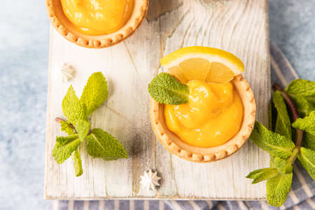 Shortbread tartlet filled with lemon curd, mint and lemon slices and mini meringue on wooden board, blue concrete background. Top view.の写真素材