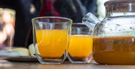 Hot spicy sea buckthorn tea in glass teapot with mint, orange and honey on wooden table in cafe....