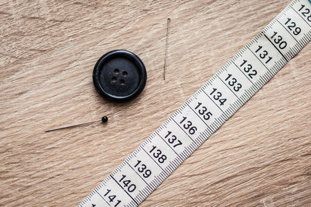 a button, two sewing needles and a measuring tape over a wooden surfaceの写真素材
