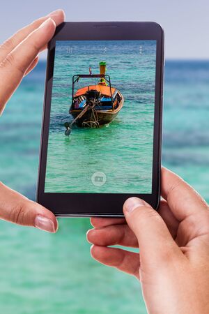 a woman using a smart phone to take a photo of a traditional thai long tail boat floating in a tropical seaの写真素材