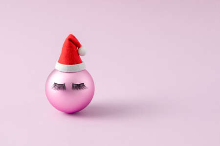 Pink Christmas bauble face with eyelashes and Santa Claus hat. Minimal creative New Year conceptの写真素材