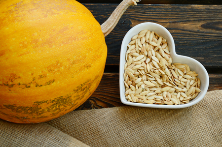 Pumpkin seeds in Heart-shaped bowl and pumpkin, sack cloth on wooden background. Health Benefits of Pumpkin Seeds.の写真素材
