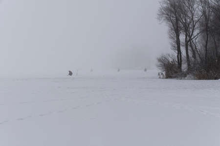 Winter fishing. Fishermen sit on the ice near the shore in a thick fog.の写真素材