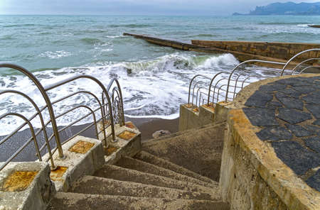 Staircase descending to the sandy beach. A strong surf reaches the lower steps. Sudak, Crimea. End of April. Out of season.の写真素材