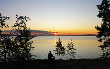 Evening at Lake Kanozero. Silhouette of a man sitting on the shore and looking at the setting sun. Umba river, Kola Peninsula, Murmansk Oblast, Russia. August.の写真素材