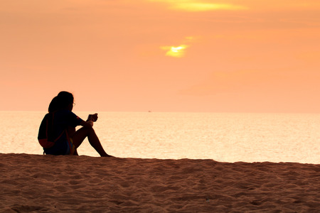 Silhouette of Girl Tourist with Backpack is Using Mobile Smart Phone on The Phuket Sea Beach with Sunset and Sea Background. Set as Blank Frame for Text.の写真素材