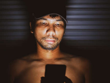 An Asian man looks at his mobile phone in a dark corner with homesickness.の写真素材
