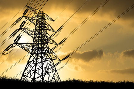 High voltage power lines and transmission tower silhouetted against a dramatic sunset sky, representing energy and infrastructure.の写真素材