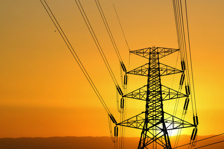 Silhouette of a high voltage power transmission tower with power lines at sunset, set against a vibrant orange sky.の写真素材