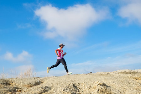 A man in athletic clothing running on a rocky trail with a clear blue sky and white clouds in the background.の写真素材