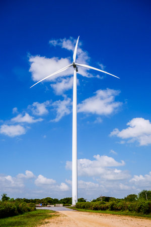 A tall wind turbine stands against a clear blue sky with white clouds, set in a rural landscape with greenery and a dirt road.の写真素材