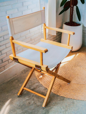 A stylish wooden director's chair in a sunlit modern interior with a white brick wall, potted plant, and woven rug.の写真素材